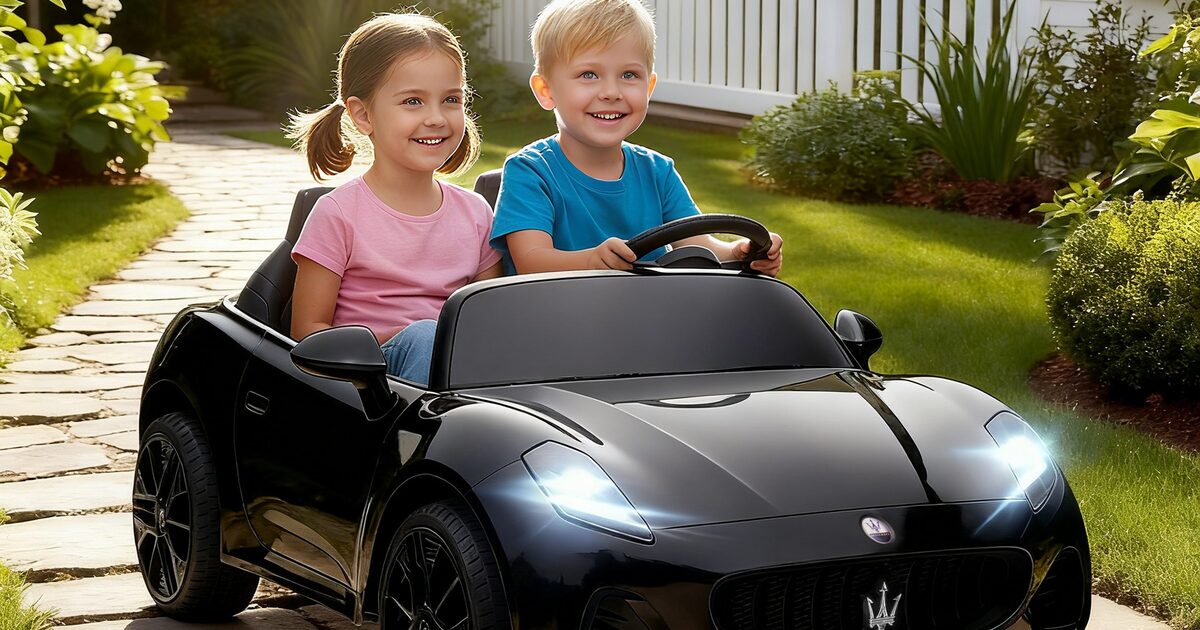 En flicka och en pojke sitter bredvid varandra i en svart tvåsitsig Maserati för barn med tända strålkastare, på en trädgårdsgång